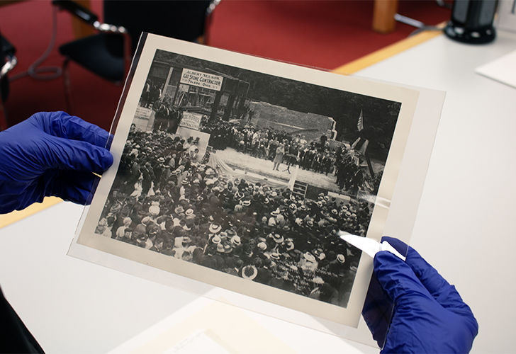 Photo of a person with blue gloves on holding a photo titled "Laying of the Corner Stone of the Alumni Memorial Building, June 17, 1908 (Photo by Lyndon)/HS671" from the image archives at the Bentley Historical Library.