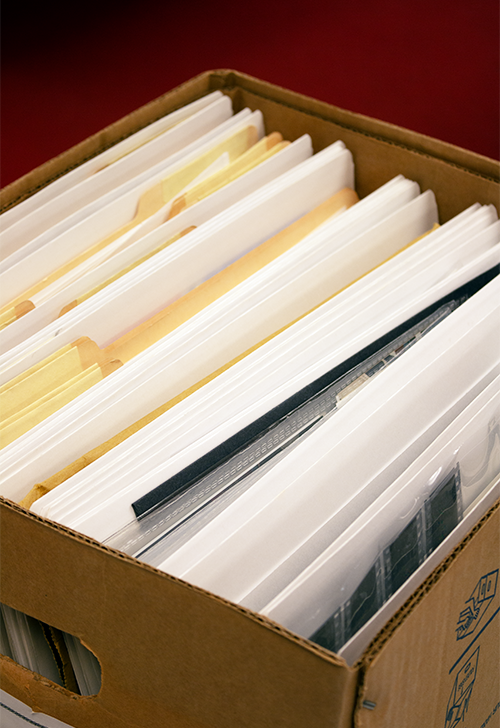 Photo of cardboard file box containing file folders and sheets of film negatives with red floor in the background taken at the Bentley Historical Library.