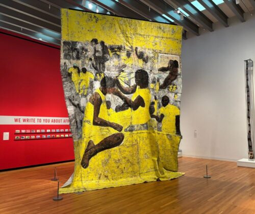A large floor-to-ceiling textile hangs from the ceiling of an art gallery. The textile is yellow, grey, and brown and depicts individuals styling others' hair.