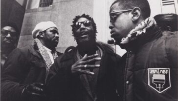 Frame from movie, “Passin’ It On” in black and white showing a group of people and one person at the center speaking.