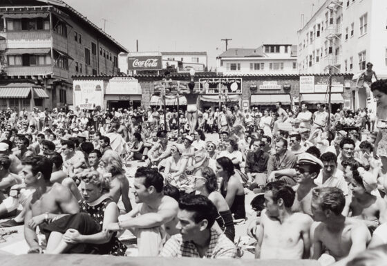 A crowd of people, many wearing bathing suits, sit viewing a platform. Behind them are storefronts and a Coca-Cola sign.