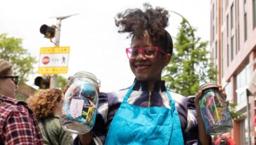African American woman with red glasses and blue apron and two jars filled with paper crafts smiling walking down a city street