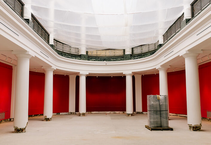 A grand, empty room under renovation. The walls are painted a bright red, and the floor is stripped down to the base. Large white columns line the sides, and natural light enters through the tall windows.