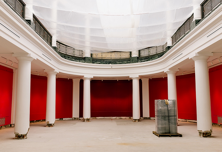 A grand, empty room under renovation. The walls are painted a bright red, and the floor is stripped down to the base. Large white columns line the sides, and natural light enters through the tall windows.