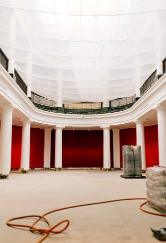 A grand, empty room under renovation. The walls are painted a bright red, and the floor is stripped down to the base. Large white columns line the sides, and natural light enters through the tall windows.