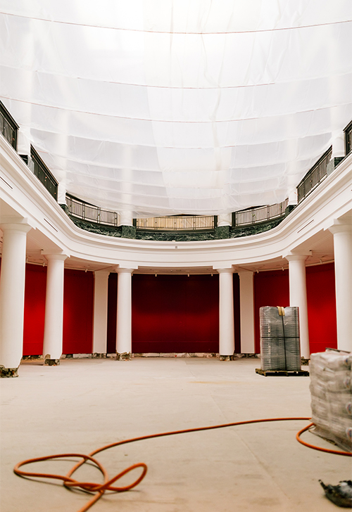 A grand, empty room under renovation. The walls are painted a bright red, and the floor is stripped down to the base. Large white columns line the sides, and natural light enters through the tall windows.