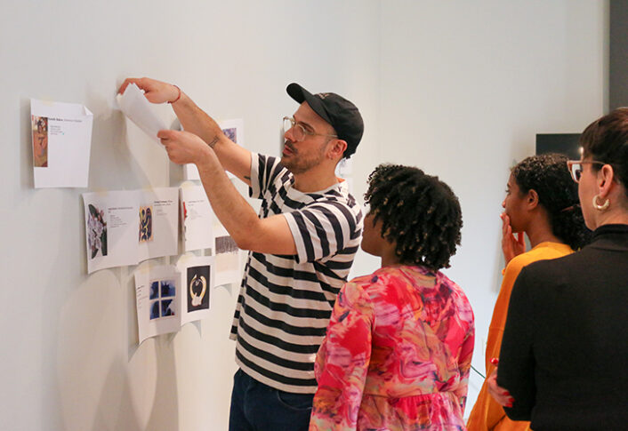 An individual taping prints of artworks on a wall while three others observe it.