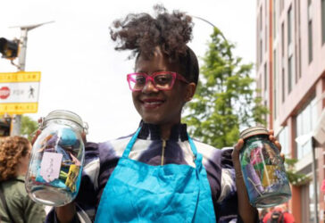 African American woman with red glasses and blue apron and two jars filled with paper crafts smiling walking down a city street