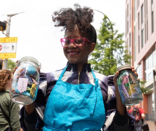 A female with Black skin and red glasses stands holding two mason jars full of colorful tags. She is wearing a bright blue apron