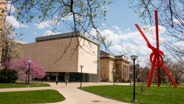 A campus scene at the University of Michigan features a large red sculpture standing prominently amidst blooming trees with pink flowers. Nearby buildings and a few people walking on a paved path can be seen in the background.