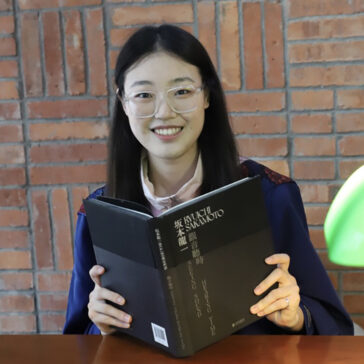 A person with long black hair and glasses holds a book and smiles at the camera