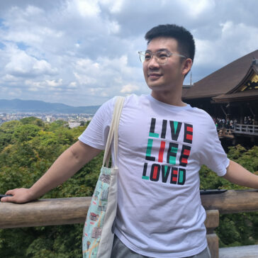 A person stand in front a lush green backdrop. His shirt read "Live Life Loved"