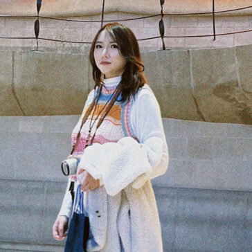 A person in a white sweater stands with a camera around her neck