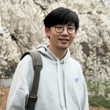 A man in a grey hooded sweatshirt stands against a background of white flowering trees
