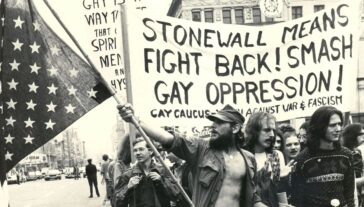 Historical image of a protest. A man holds an american flag and a banner behind him reads "Stonewall means fight back!"
